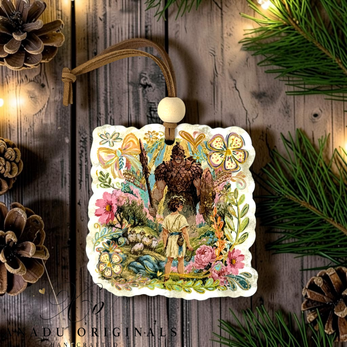 A decorative square ornament with scalloped edges featuring an illustrated David and Goliath featuring a colorful folk art storybook illustration of young David facing the giant among vibrant floral meadows.. The ornament hangs from a brown cord with a white bead, displayed on a rustic wood surface with pine branches, pinecones, and warm string lights.