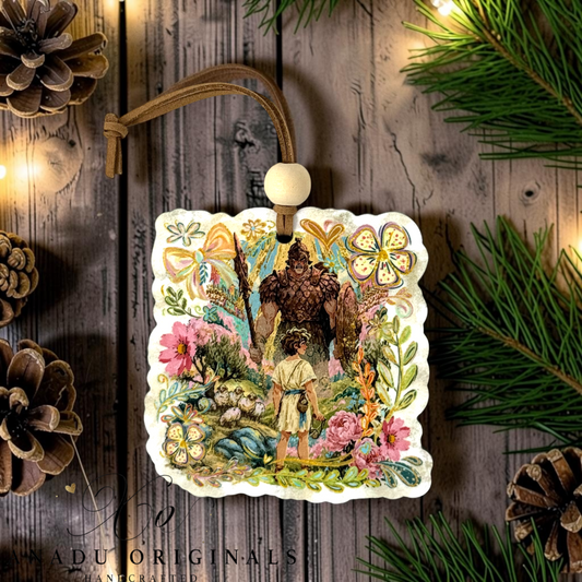 A decorative square ornament with scalloped edges featuring an illustrated David and Goliath featuring a colorful folk art storybook illustration of young David facing the giant among vibrant floral meadows.. The ornament hangs from a brown cord with a white bead, displayed on a rustic wood surface with pine branches, pinecones, and warm string lights.