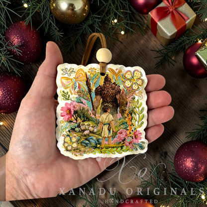 A hand holds a square, scalloped Retro religious Bible story Christmas ornament featuring an illustrated scene of a boy and a man in a forest, surrounded by colorful floral and bow motifs. The ornament is displayed against a festive holiday background with pine branches, red ornaments, and a gift box.