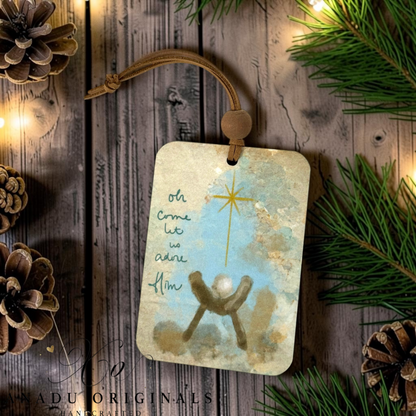 Oh Come Let Us Adore Him wood ornament showing a rustic baby Jesus manger under a large gold star on a textured blue and gold watercolor background.
