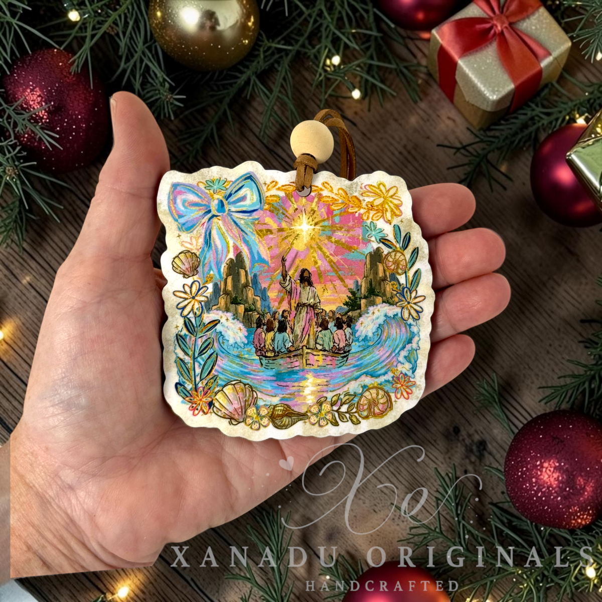 A hand holds a handcrafted Christmas ornament featuring a colorful illustration of Jesus and men in a boat, surrounded by decorative pine branches, ornaments, and gift boxes. The ornament has a scalloped edge and a hanging loop at the top.