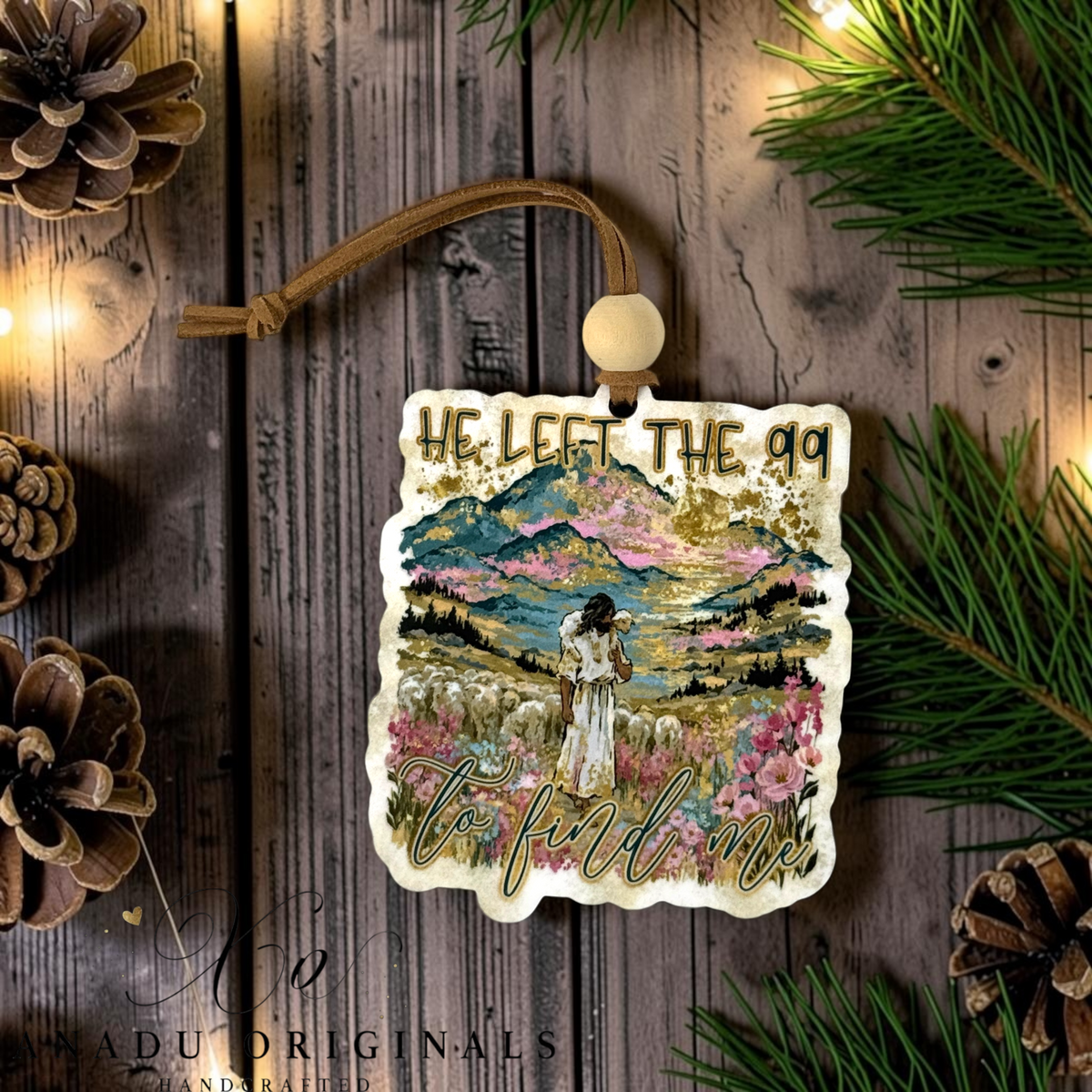 A Christmas ornament featuring a colorful illustration of Jesus leading a flock of sheep through a field, with the text 'HE LEFT THE 99 TO FIND ME' in gold lettering. The ornament is displayed on a rustic wooden background decorated with pine branches, pinecones, and warm string lights.