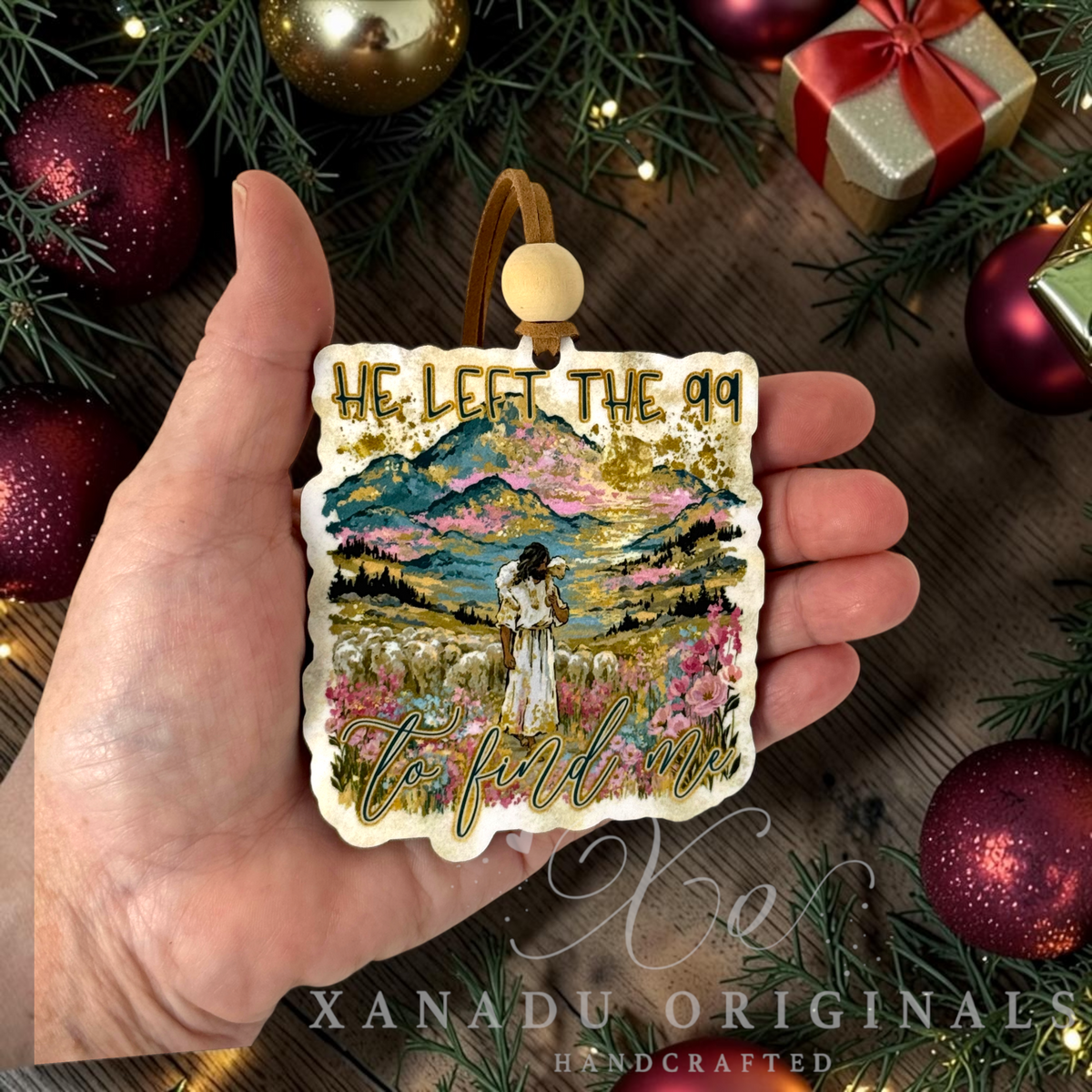 A hand holds a square decorative ornament featuring a colorful mountain landscape illustration with Jesus in a field and gold glitter text reading 'HE LEFT THE 99 TO FIND ME.' The ornament is shown against a holiday-themed background with ornaments, evergreen branches, and a wrapped gift.