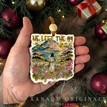 A hand holds a square decorative ornament featuring a colorful mountain landscape illustration with Jesus in a field and gold glitter text reading 'HE LEFT THE 99 TO FIND ME.' The ornament is shown against a holiday-themed background with ornaments, evergreen branches, and a wrapped gift.