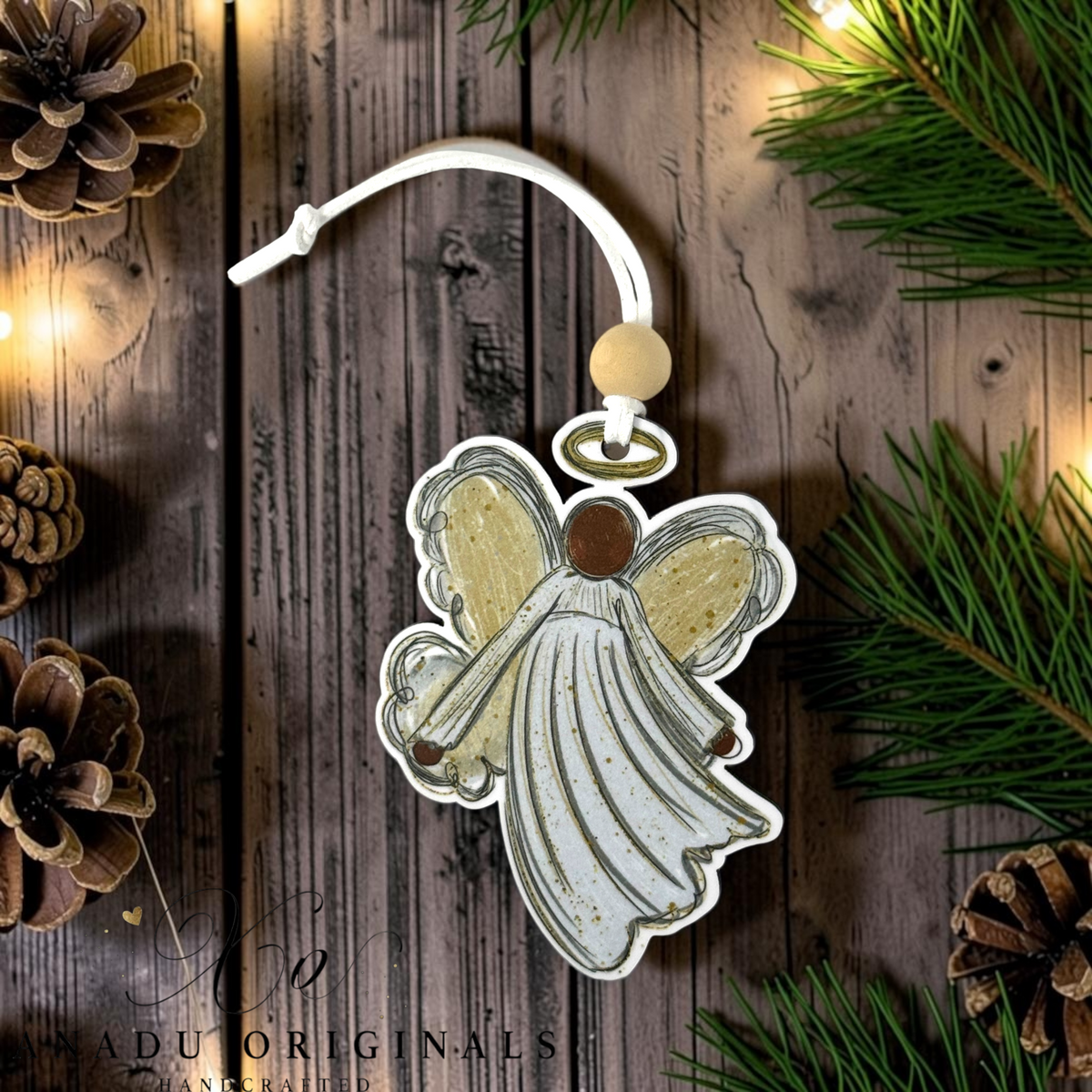 A handcrafted Christmas angel ornament with distressed white wings and a white robe, hanging from a white cord with a wooden bead. The ornament is displayed on a rustic dark wood background surrounded by pine needles, pinecones, and warm string lights.