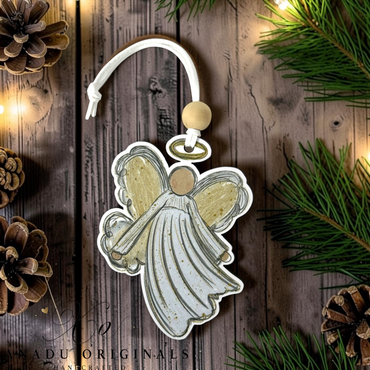 A Christmas angel ornament with a white and gold distressed design, hanging from a white cord with a wooden bead and gold ring, displayed on a rustic wooden background with pine needles and pinecones.