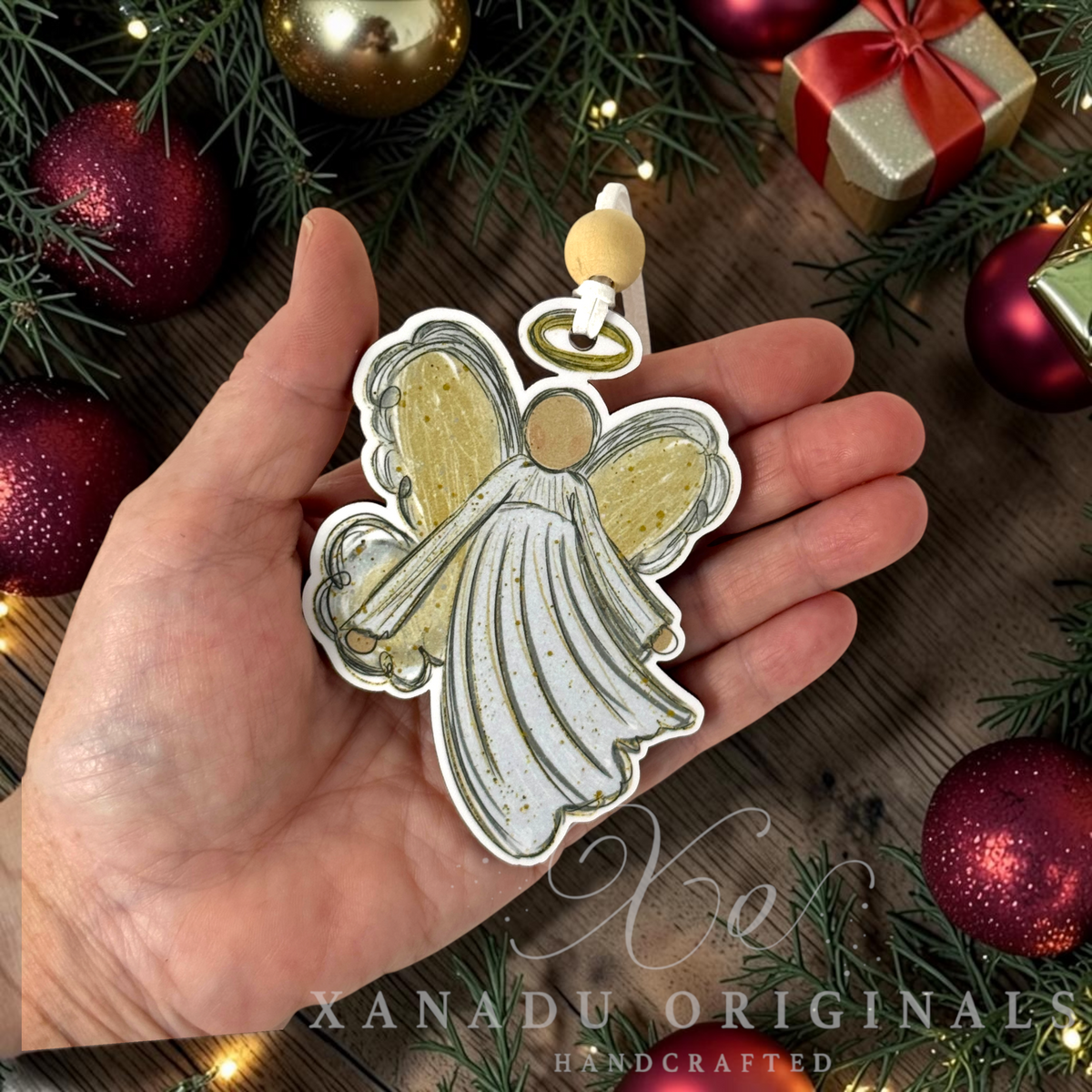 A hand holds a handcrafted Christmas angel ornament by Xanadu Originals. The ornament features a white and gold glittery angel design with a gold-colored wooden bead loop at the top.