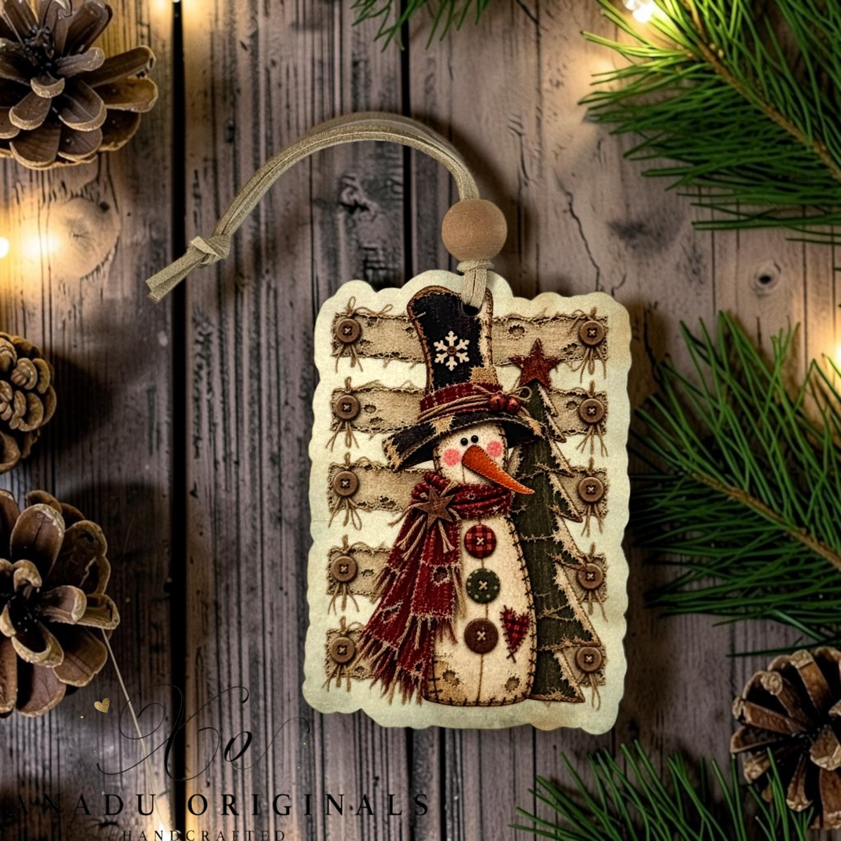 A handmade Christmas ornament featuring a stitched snowman wearing a black top hat and red scarf, hanging from a cord on a rustic wooden surface surrounded by pine branches, pinecones, and warm string lights. The brand name 'ADU ORIGINALS' is visible in the bottom left corner.