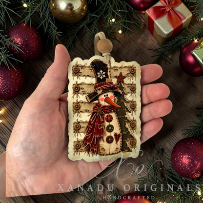 A hand holds a rectangular handmade Christmas ornament featuring a stitched snowman wearing a black top hat and red scarf, surrounded by pine branches, red ornaments, and a gold gift box. The ornament has scalloped edges and a hanging loop.