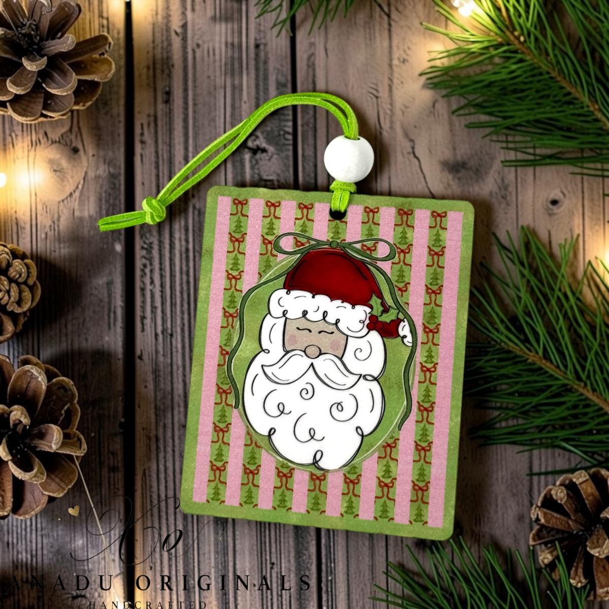 A square Christmas ornament with a green cord and white bead, featuring a cartoon Santa face on pink and green striped wood, styled on a rustic wood background with pine branches and pinecones.