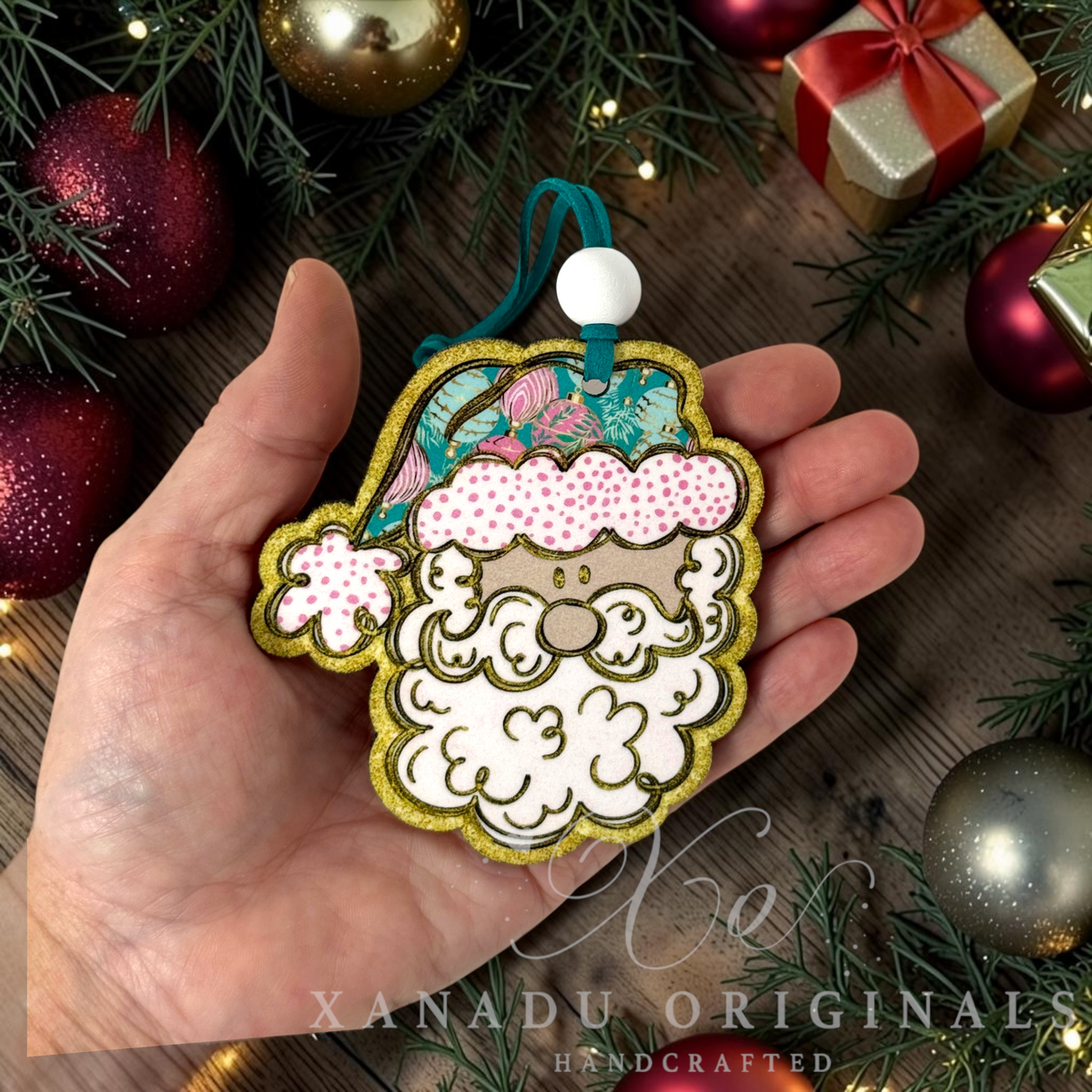 Whimsical retro pink and teal Santa Christmas ornament measuring 3.44 by 3.75 inches held in a hand next to holiday decorations.
