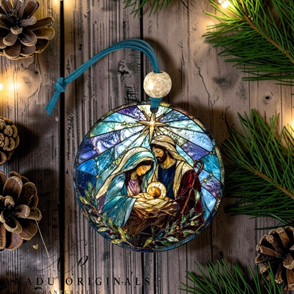 A round stained-glass style Christmas ornament featuring Mary, Joseph, and baby Jesus in a manger, hanging from a teal cord with a white bead. The ornament is displayed on a rustic wooden surface surrounded by pine branches, pinecones, and warm string lights.