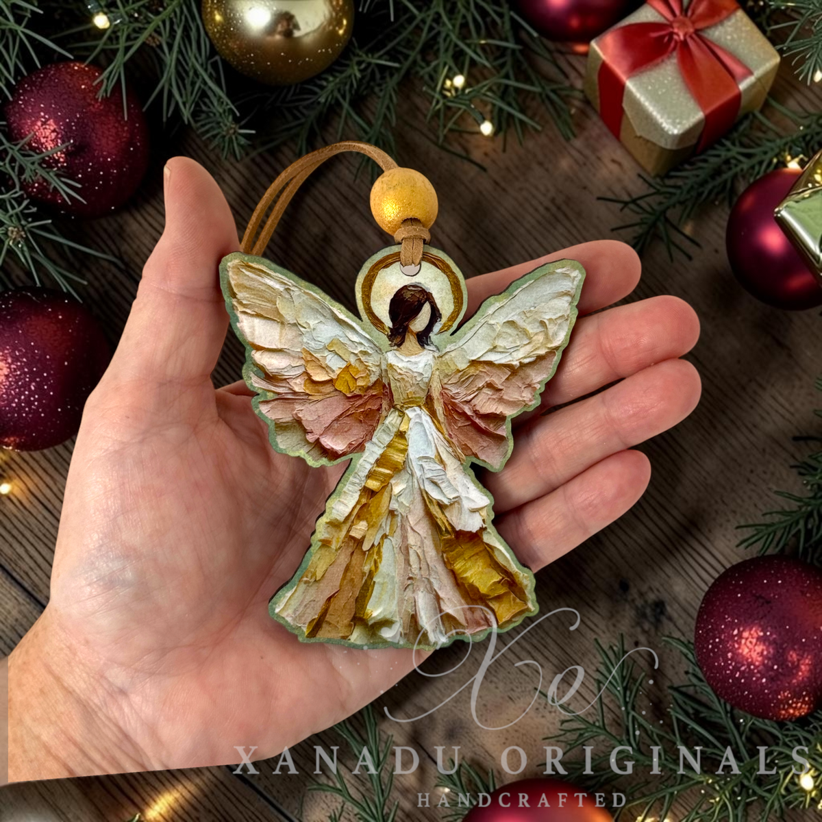 A hand holds a handcrafted Christmas angel ornament with textured, layered wings in cream, gold, and muted pink, set against a rustic holiday background with pine branches, red ornaments, and a gold gift box. A watermark at the bottom reads 'XANADU ORIGINALS HANDCRAFTED'.