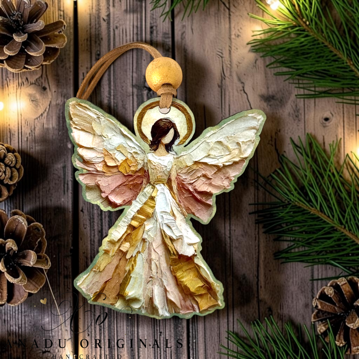 An angel-shaped Christmas ornament with textured, layered wings in cream, gold, and dusty pink tones, hanging from a brown cord with a wooden bead. The ornament is displayed on a rustic wooden background decorated with pine needles, pinecones, and warm string lights.