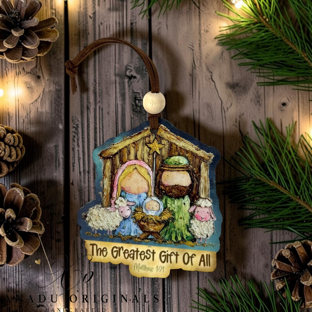 A handcrafted wooden Christmas ornament featuring a nativity scene with Mary, Joseph, baby Jesus, animals, and a star, hanging from a brown leather loop with a white bead. The ornament has a decorative text plaque at the bottom reading 'The Greatest Gift Of All (Matthew 1:2)'.