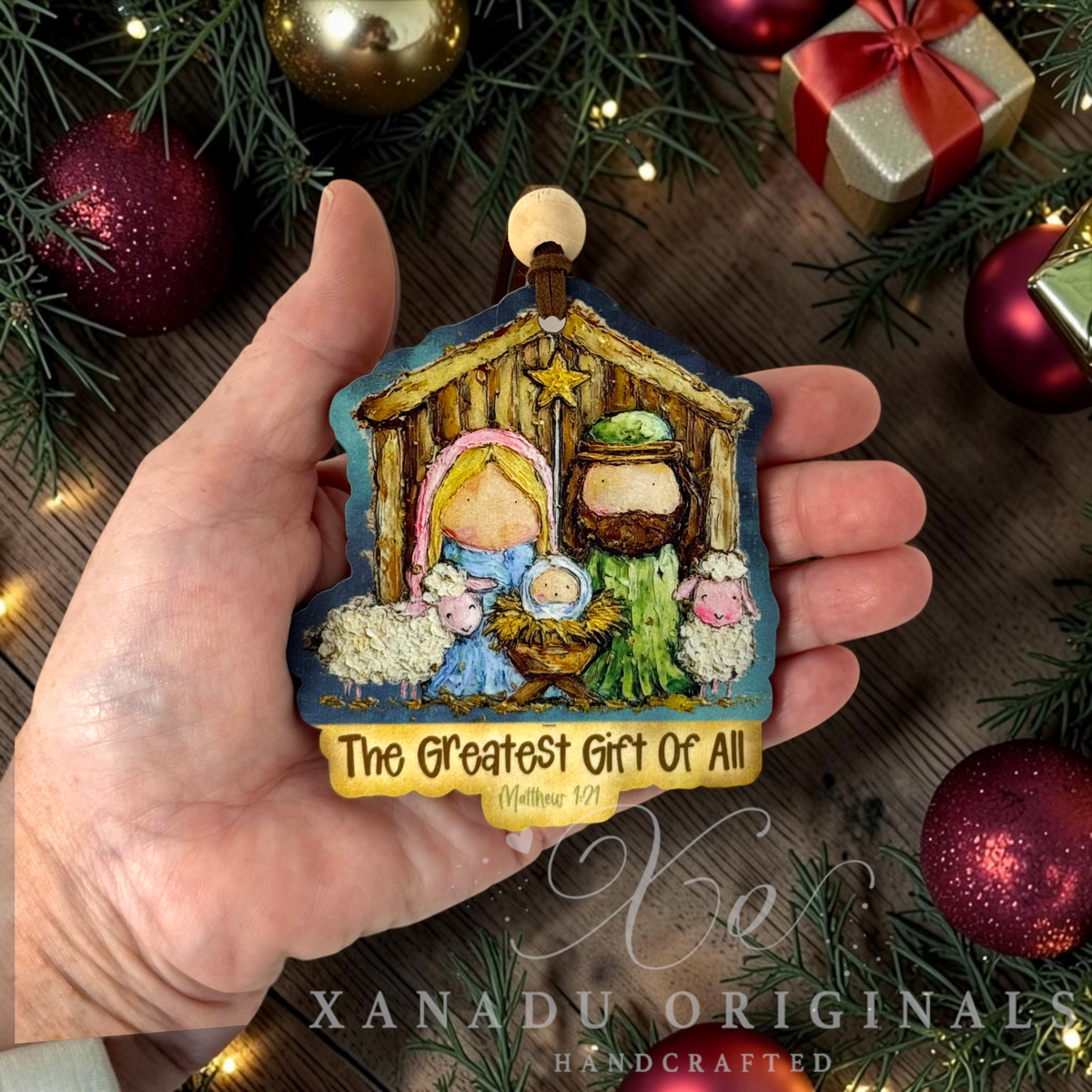 A hand holds a handcrafted Christmas ornament shaped like a stable with Mary, Joseph, baby Jesus, and sheep. The ornament has a gold hanging loop and a gold banner reading 'The Greatest Gift Of All' with 'Matthew 1:21 below.