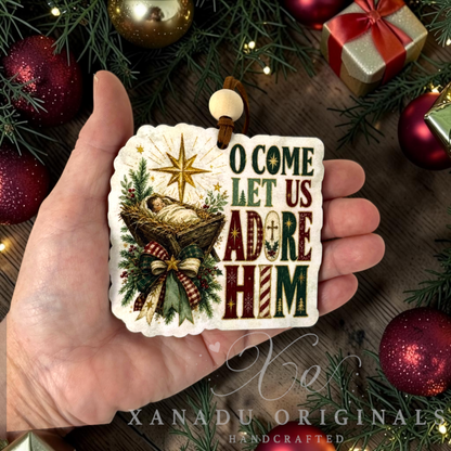 A hand holds a handcrafted Christmas ornament featuring the text 'O COME LET US ADORE HIM' with a nativity scene and gold star. The background is decorated with pine branches, red ornaments, and a gift box.