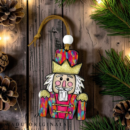 A hand-painted wooden Christmas nutcracker ornament with a colorful patterned coat, gold crown, and white beard, hanging on a rustic wooden background with pine branches and warm string lights.