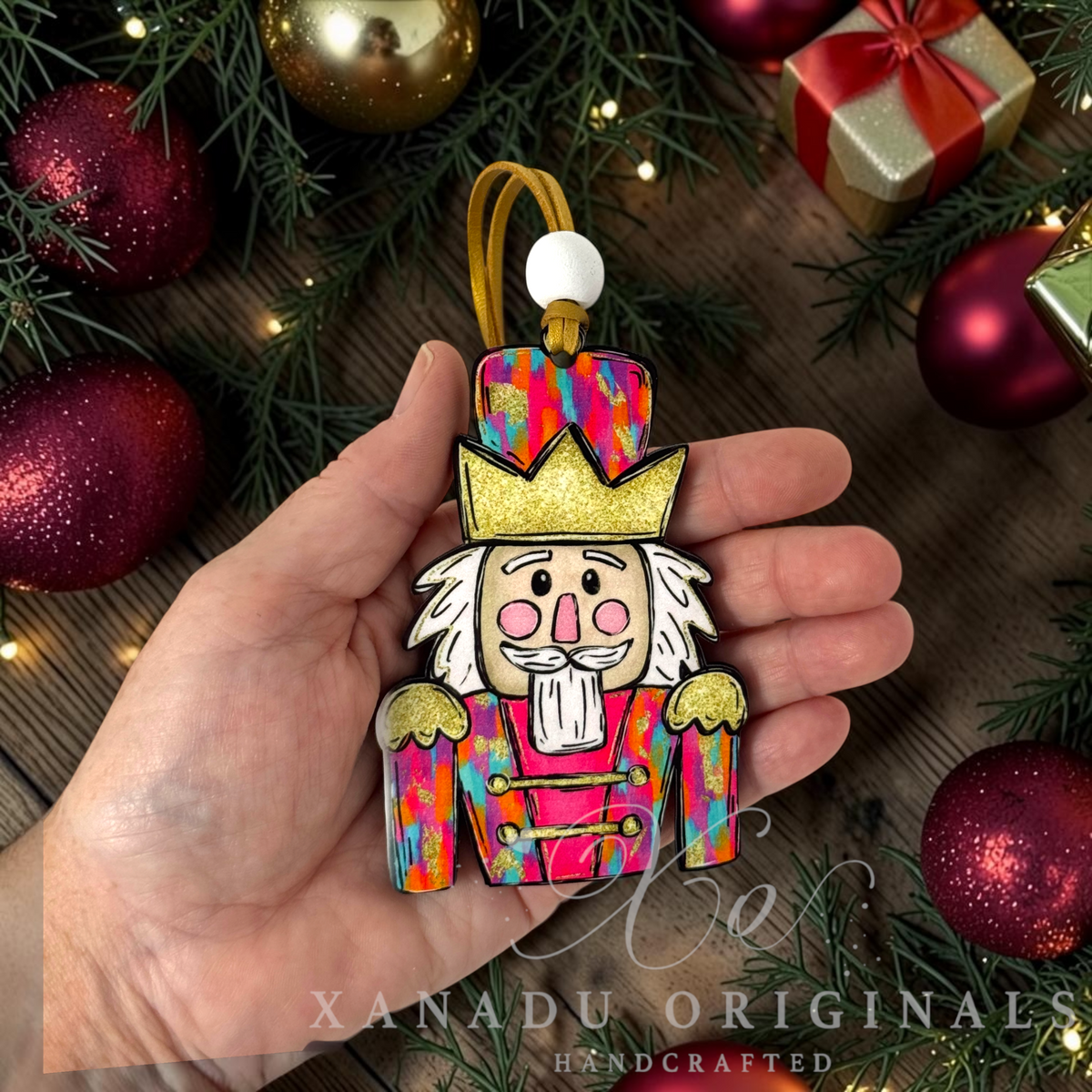 A hand holds a colorful nutcracker ornament decorated with a vibrant multicolor pattern, gold crown, and white beard, surrounded by Christmas decorations including red ornaments, pine branches, and a gift box.