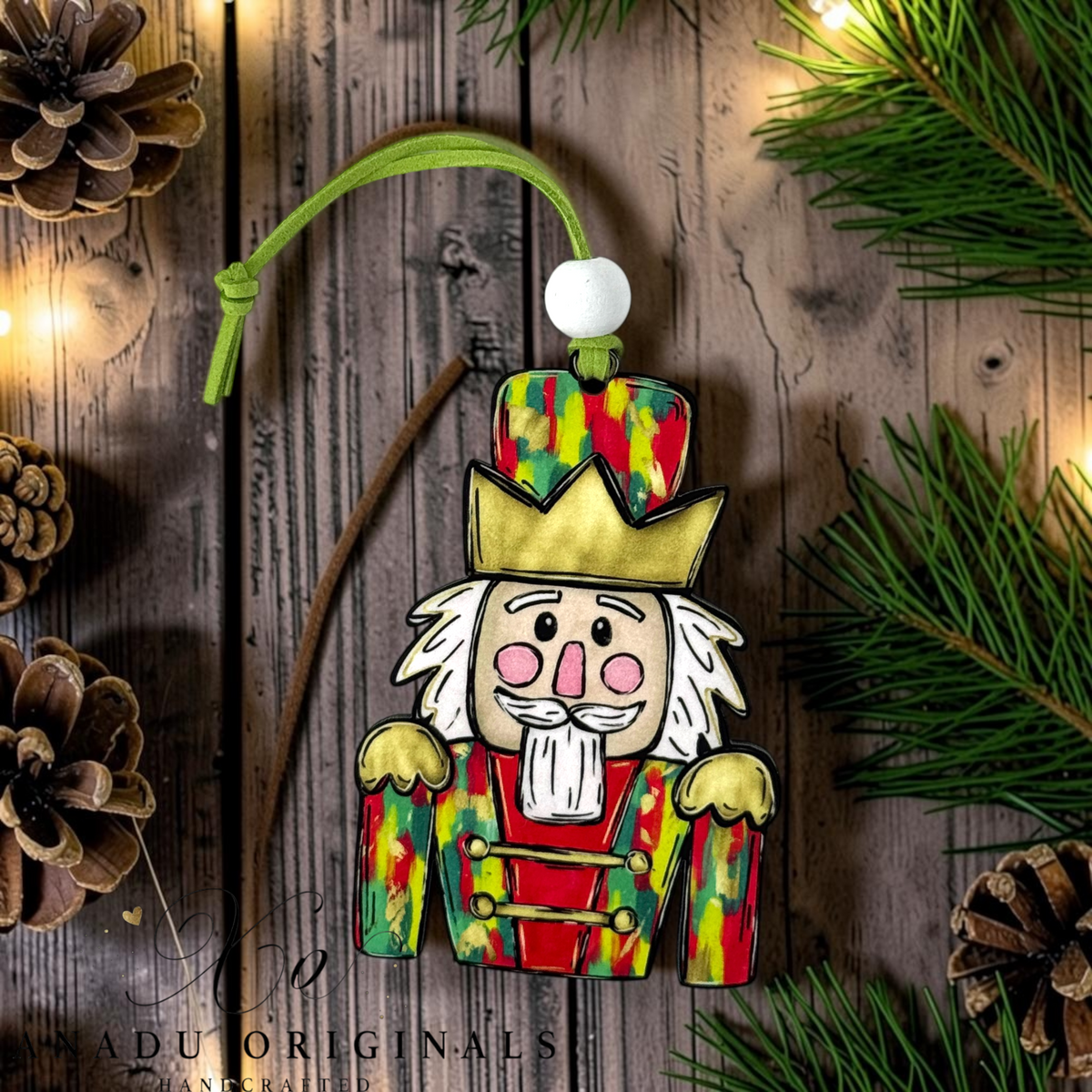 A handcrafted wooden Christmas nutcracker ornament with a colorful paint-splattered red and green uniform, gold crown, white beard, and pink cheeks, hanging on a rustic wood background with pine branches, pinecones, and warm string lights.