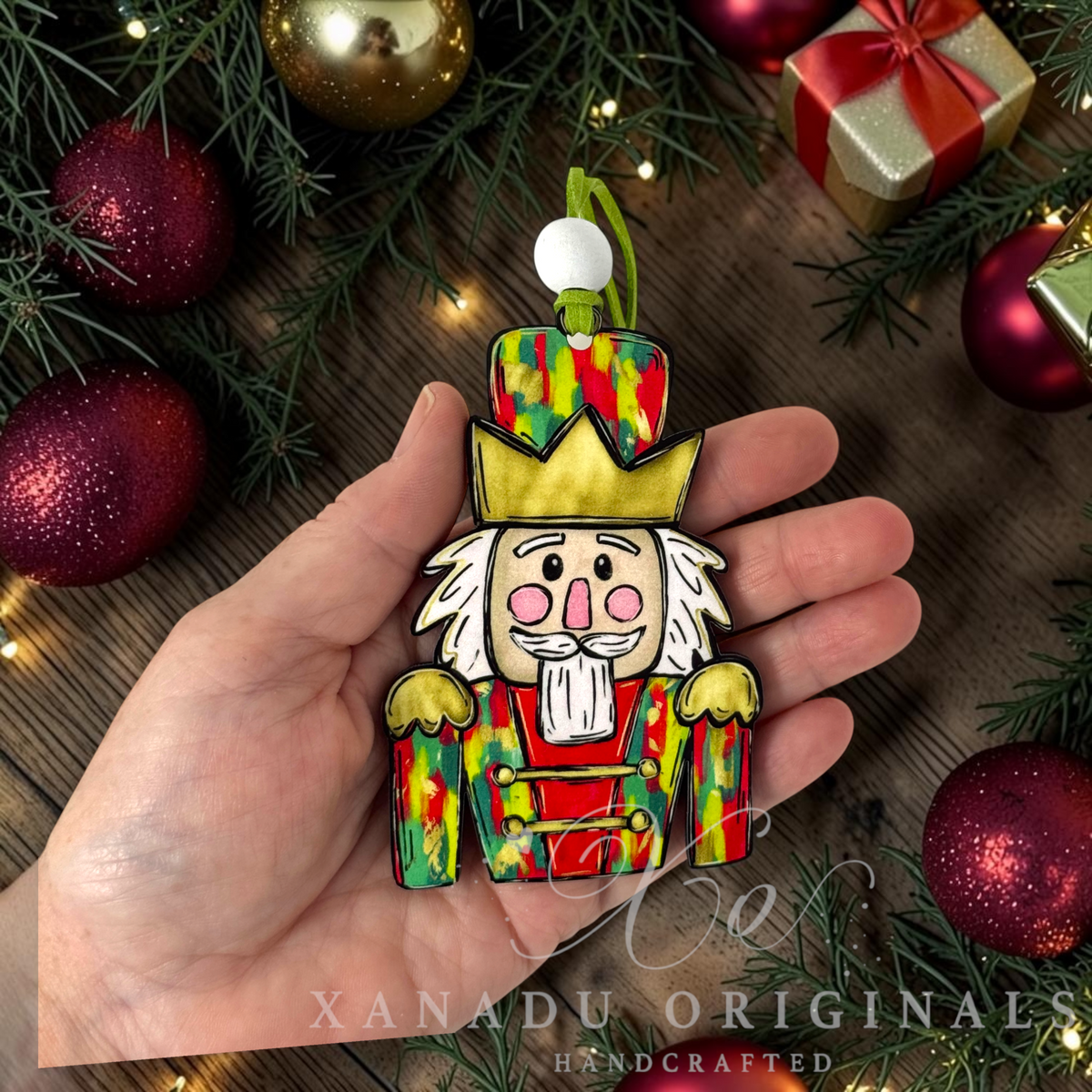 A hand holds a colorful handcrafted nutcracker ornament against a rustic wooden background decorated with pine branches, red ornaments, and a gold gift box. The ornament features a green and red patterned hat, a gold crown, and a white beard.