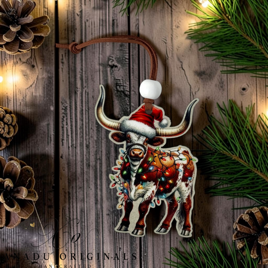 A decorative hanging ornament shaped like a longhorn bull wearing a Santa hat, with a colorful holiday wreath and warm lights, shown on a rustic wood background with pine branches and pinecones.