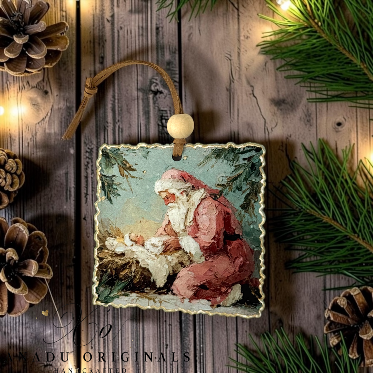 A square Christmas ornament featuring a painted scene of Santa Claus kneeling by a baby in a snowy setting, hanging from a brown cord with a wooden bead. The ornament is styled on a rustic wooden surface with pine branches, pinecones, and warm string lights.