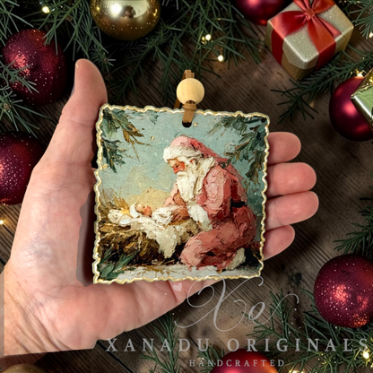 A hand holds a square scalloped-edge Christmas ornament featuring an oil painting of Santa Claus cradling baby Jesus. The ornament is decorated with a gold-toned border and a light wood hanging loop.