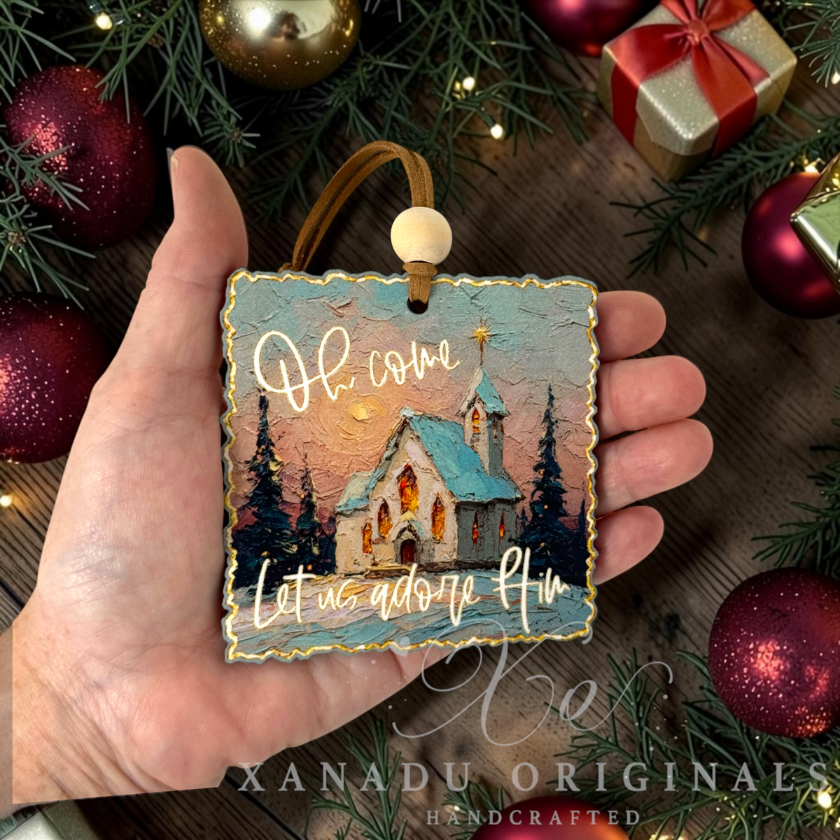 A hand holds a square Christmas ornament featuring a painted church scene with the words 'Oh come Let us adore Him' in gold lettering. The ornament has a gold border, a tan hanging cord, and a white bead.