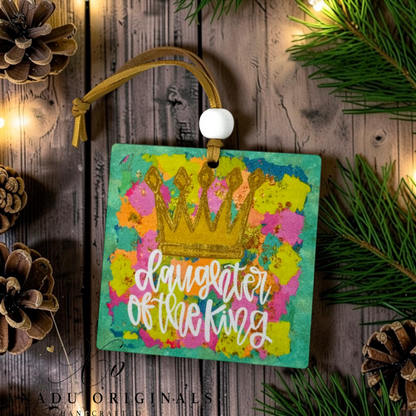 A square decorative hanging ornament with a colorful painted background, a gold crown graphic, and the phrase “daughter of the king” in white script. It hangs from a tan cord with a white bead, styled on a rustic wood surface with pine branches, pinecones, and warm string lights.