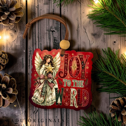 A decorative Christmas ornament featuring an angel illustration and the words “JOY TO THE WORD” on a red background, hanging from a brown cord with a wooden bead. The ornament is styled on a rustic wood surface with pine branches, pinecones, and warm string lights.