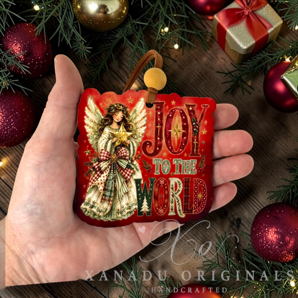 A hand holds a red retro Christmas ornament featuring an angel and the words 'JOY TO THE WORD' in decorative lettering. The scene is styled with pine branches, red ornaments, a gold bauble, and a wrapped gift, with a watermark reading 'Xanadu Originals Handcrafted' at the bottom.