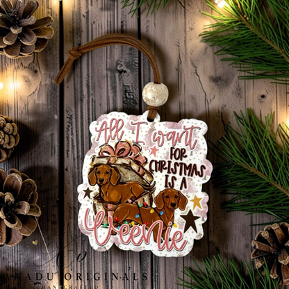 A die-cut Christmas ornament featuring an illustration of two brown dachshunds with a gift and the text 'All I want for Christmas is a Weenie', hanging from a brown cord on a rustic wooden background with pine branches and pinecones.