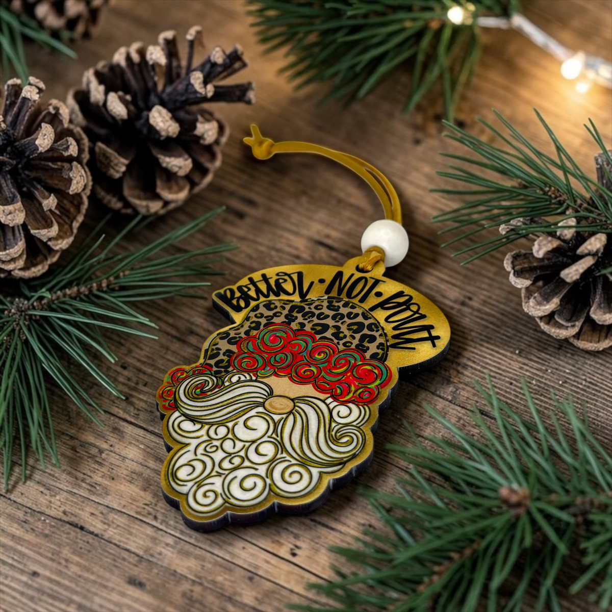 A decorative Christmas ornament shaped like a stylized Santa face with a gold border, leopard-print forehead, red and cream swirl beard, and the words “Better-NOT-Pout” in black script. The ornament hangs from a gold string with a white bead and is photographed on a rustic wooden surface surrounded by pine needles and pinecones.