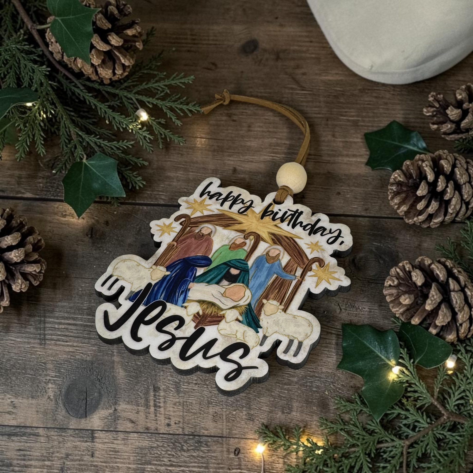 A decorative wooden Christmas ornament shaped like a nativity scene with the words 'happy birthday, Jesus' is displayed on a rustic wooden surface surrounded by pinecones, evergreen branches, and warm fairy lights.