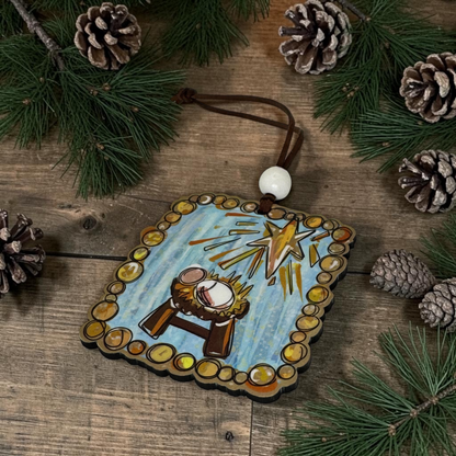 A square wooden Christmas ornament with a nativity scene, featuring a baby in a manger under a shining star, hanging from a brown cord with a white bead. The ornament is displayed on a rustic wooden surface surrounded by pine branches and pinecones.
