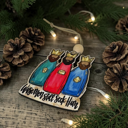 A hand-painted wooden Christmas ornament depicting the Three Wise Men with the text 'Wise Men still seek Him' on a rustic wooden surface surrounded by pinecones and evergreen branches with warm string lights.