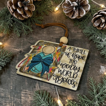 A wooden Christmas ornament featuring an angel illustration and the quote 'A THRILL of HOPE THE HEAVY WORLD WILL FALL' on a rustic dark wood background with pine branches, pinecones, and warm string lights.