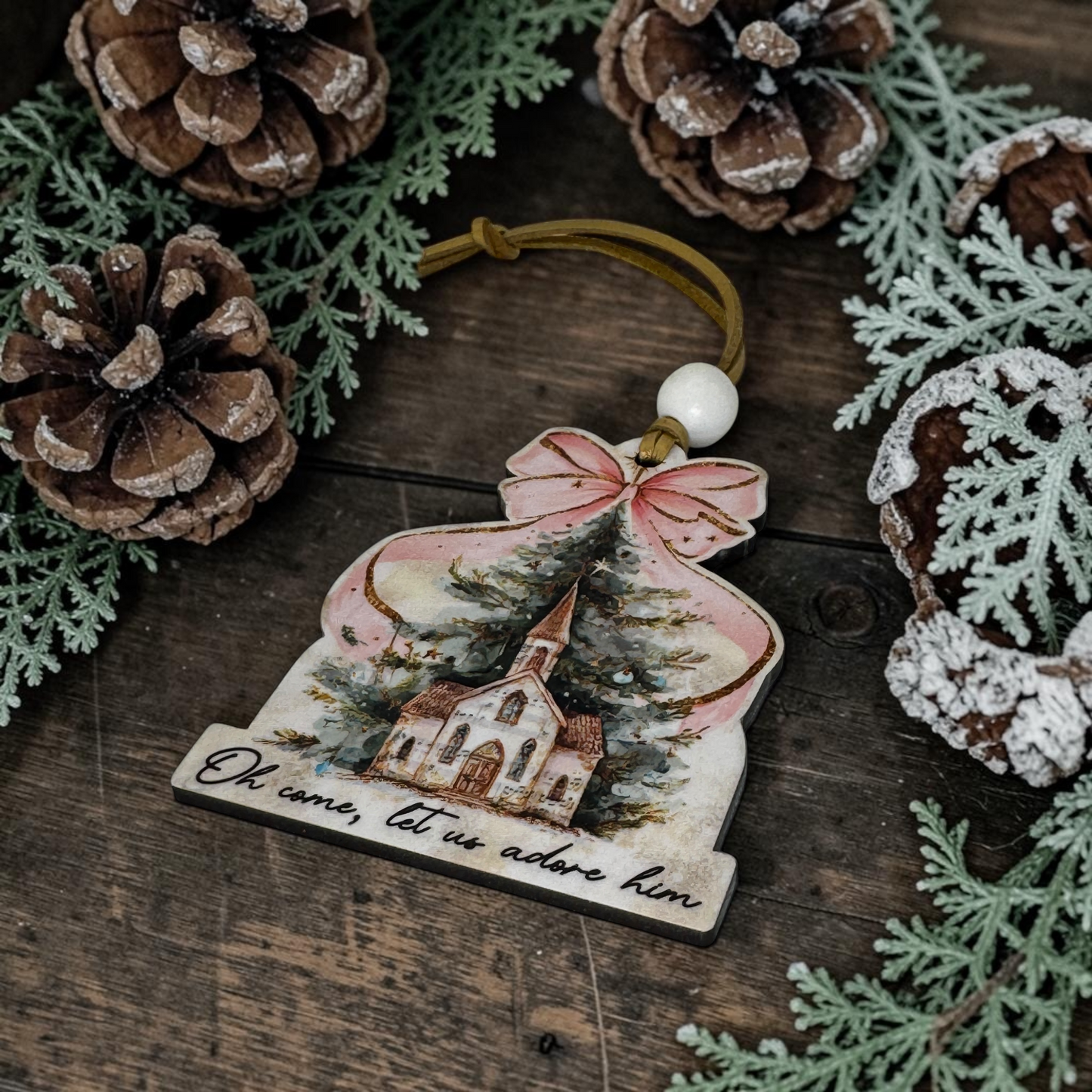 A hand-painted Christmas ornament shaped like a bell, featuring a church scene with the text 'Oh come, let us adore him' and a hanging loop, styled on a rustic wooden surface with pinecones and greenery.