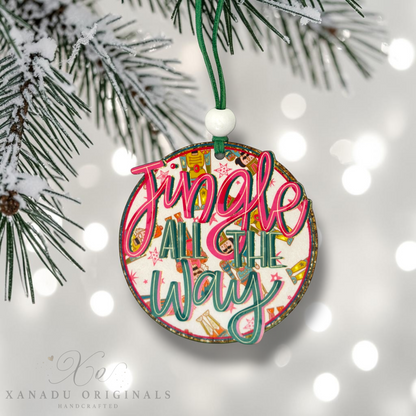 A round wooden Christmas ornament hanging from a green pine branch. The ornament features the phrase 'Jingle All The Way' in pink and teal script over a colorful holiday pattern, with a white bead and green hanging cord above it.