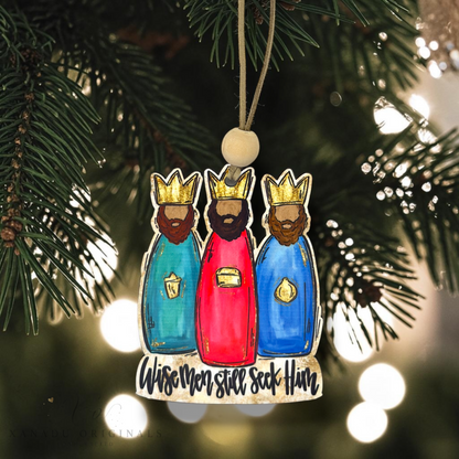 A hand-painted wooden Christmas ornament depicting the Three Wise Men, hanging from a pine tree branch with warm bokeh lights in the background. The ornament features gold crowns, detailed robes in teal, red, and blue, and the text 'Wise men still seek Him' at the bottom.