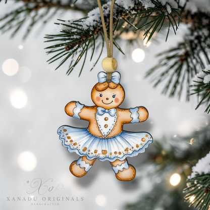 A handcrafted gingerbread girl ballerina Christmas ornament hanging from a snowy pine branch with warm bokeh lights in the background. The ornament features a brown gingerbread figure with a blue bow, white and blue tutu, and gold accents.