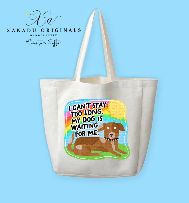 A cream-colored polyester linen tote bag featuring a vibrant cartoon graphic with a retro rainbow backdrop. In the foreground, a brown dog with white paws and a studded collar lies on green grass beneath bold black text reading "I CAN'T STAY TOO LONG. MY DOG IS WAITING FOR ME."
