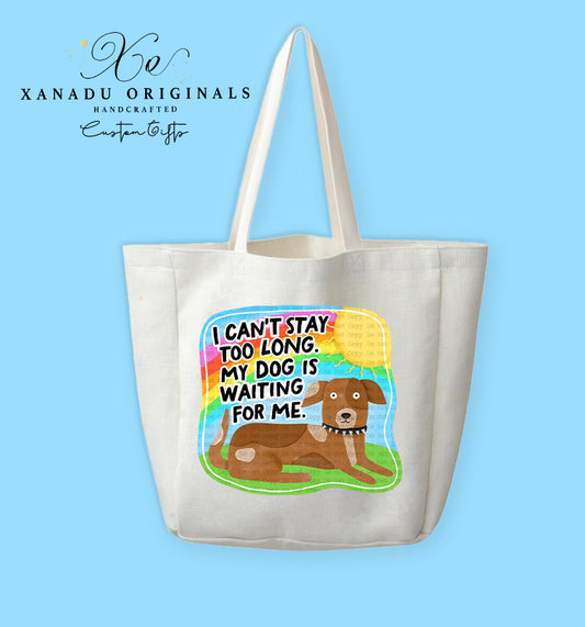 A cream-colored polyester linen tote bag featuring a vibrant cartoon graphic with a retro rainbow backdrop. In the foreground, a brown dog with white paws and a studded collar lies on green grass beneath bold black text reading "I CAN'T STAY TOO LONG. MY DOG IS WAITING FOR ME."