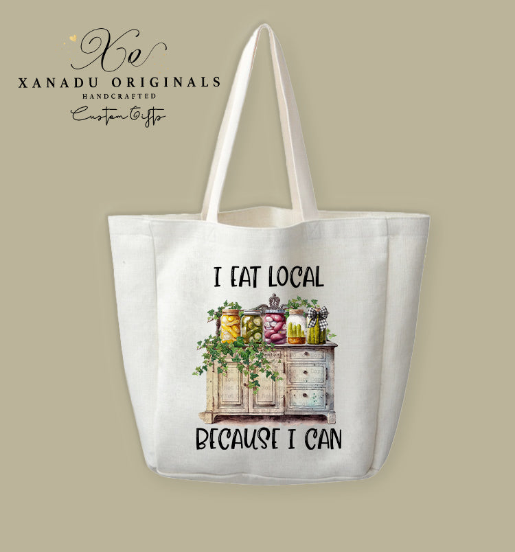 A cream-colored polyester linen tote bag featuring a watercolor illustration of a rustic white cabinet holding several colorful glass jars of canned goods, accented with green ivy vines and a black checkered bow, under black text reading "I EAT LOCAL BECAUSE I CAN."
