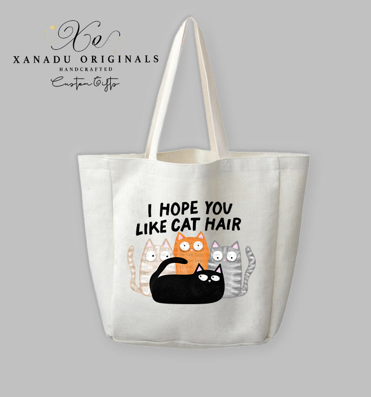 A cream-colored polyester linen tote bag featuring a cartoon illustration of four wide-eyed cats (black, orange, grey striped, and beige patterned) grouped together beneath bold black text that reads "I HOPE YOU LIKE CAT HAIR."
