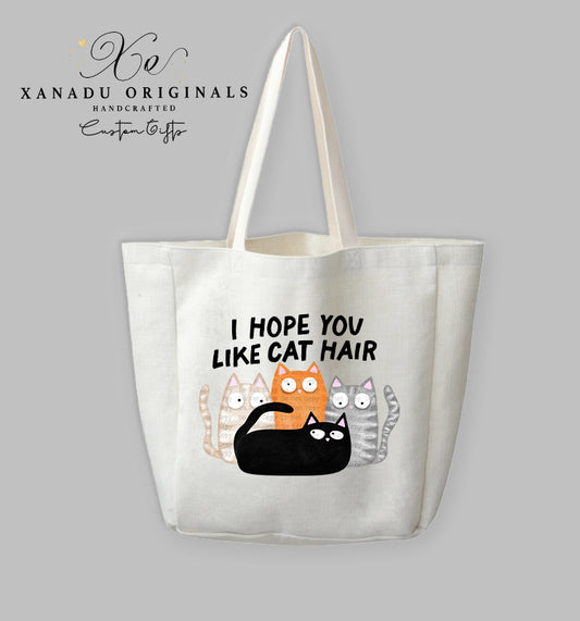 A cream-colored polyester linen tote bag featuring a cartoon illustration of four wide-eyed cats (black, orange, grey striped, and beige patterned) grouped together beneath bold black text that reads "I HOPE YOU LIKE CAT HAIR."