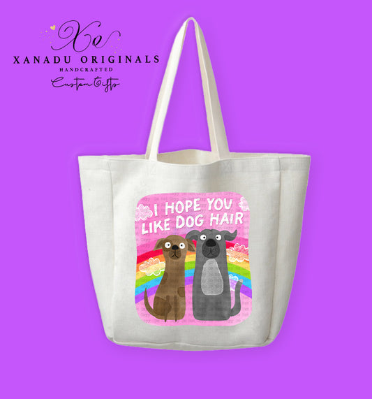 A cream-colored polyester linen tote bag featuring a square pink graphic with a retro rainbow and white clouds. In front sit two wide-eyed cartoon dogs (one brown, one grey) below bold white text that reads "I HOPE YOU LIKE DOG HAIR."