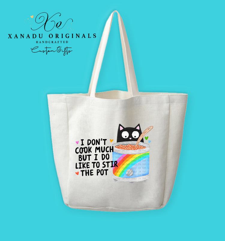 A cream-colored polyester linen tote bag featuring a cartoon illustration of a wide-eyed black cat stirring a wooden spoon inside a grey stockpot wrapped with a retro rainbow stripe. To the left, bold black text reads "I DON'T COOK MUCH BUT I DO LIKE TO STIR THE POT" surrounded by small, colorful hearts.