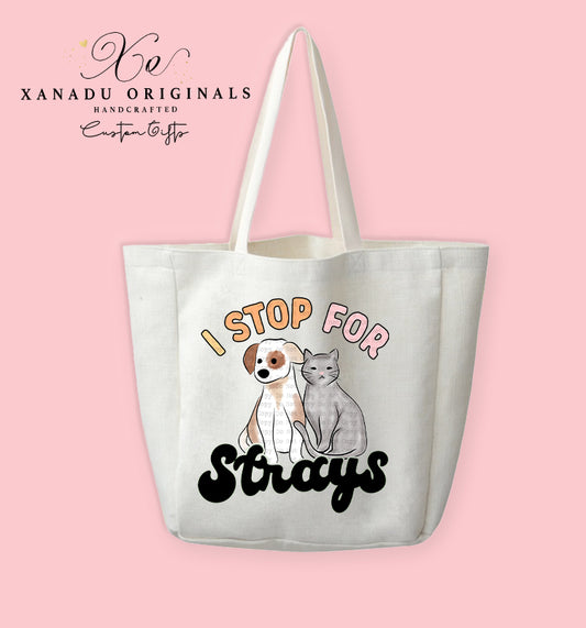 A cream-colored polyester linen tote bag featuring a watercolor illustration of a white puppy with brown spots sitting next to a grey cat. Peach bubble letters above read "I STOP FOR" and a bold black retro font below reads "Strays."